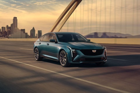 A teal Cadillac sedan drives on a highway, with a city skyline and sunset in the background.